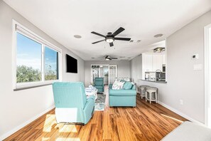 A spacious living room with a large window, two sofas, a ceiling fan, and a kitchen area in the background. - Redicare Resort - Upstairs (Galveston)
