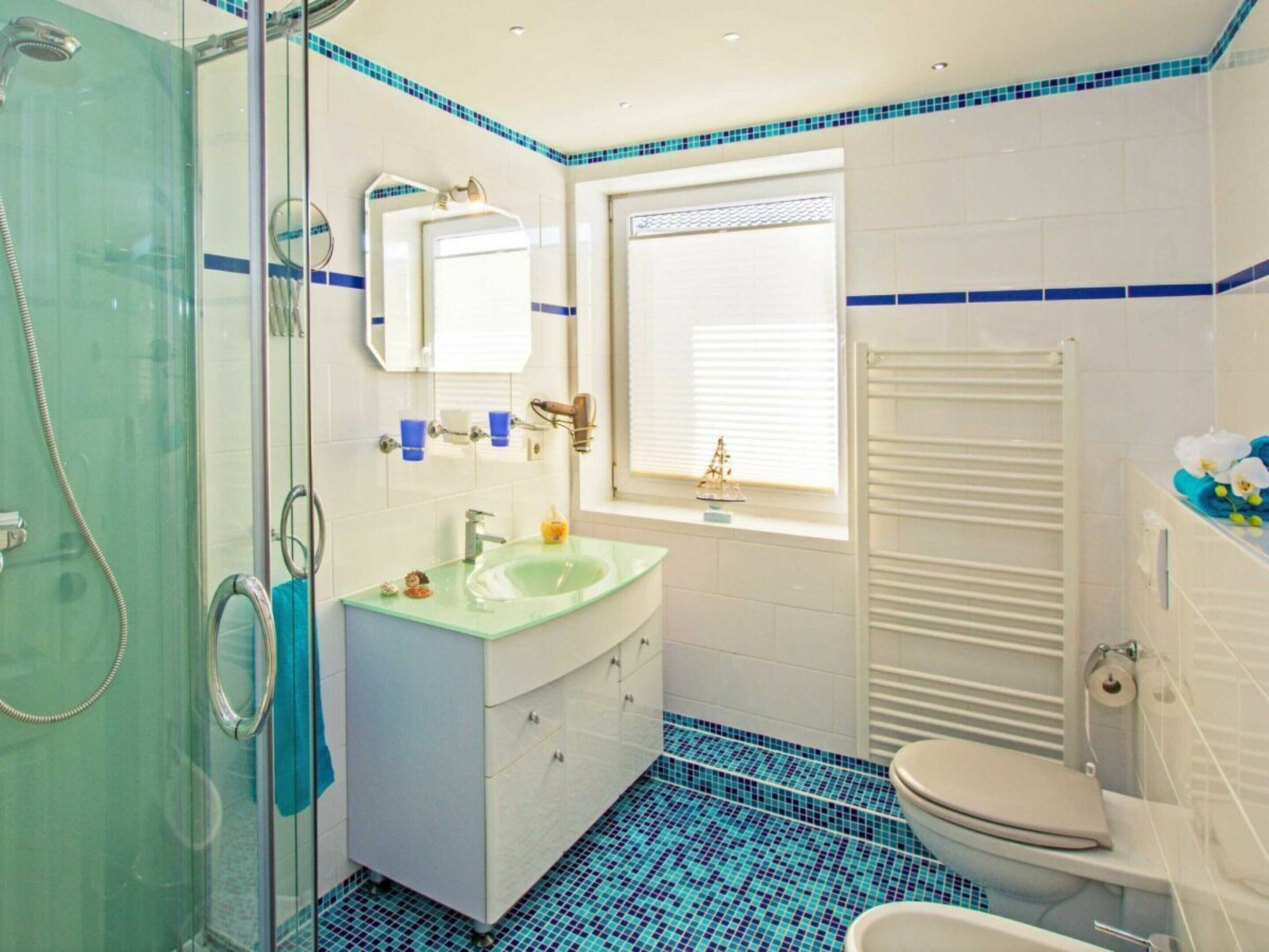 A bathroom with a glass shower enclosure, a sink with a mirror, a toilet, and a window with blinds.