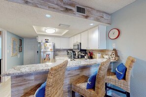 A kitchen with a central island, white cabinets, and a microwave. There are wicker chairs with blue cushions. - Surfside II Unit 309 (South Padre Island)