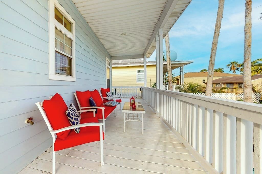 A covered porch with white railings, red and white outdoor furniture, and a view of palm trees and buildings in the background.