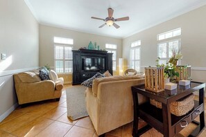 A living room with two beige sofas, a black entertainment unit, a ceiling fan, and a window with blinds. - Lantana Oasis - Private Pool, Hot Tub & Dogs OK (South Padre Island)