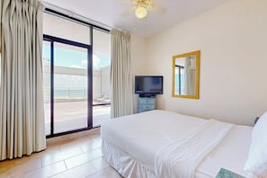 A hotel room with a large bed, a flat-screen TV, a ceiling fan, and a view through a sliding glass door. - Sunchase IV #715 - Gulf Views, Pools & Hot Tub (South Padre Island)