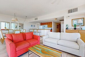 Ein Wohnzimmer mit einem roten Sofa, einem weißen Sofa und einem Couchtisch. Im Hintergrund ist eine Küche mit Holzschränken und einer Mikrowelle zu sehen.