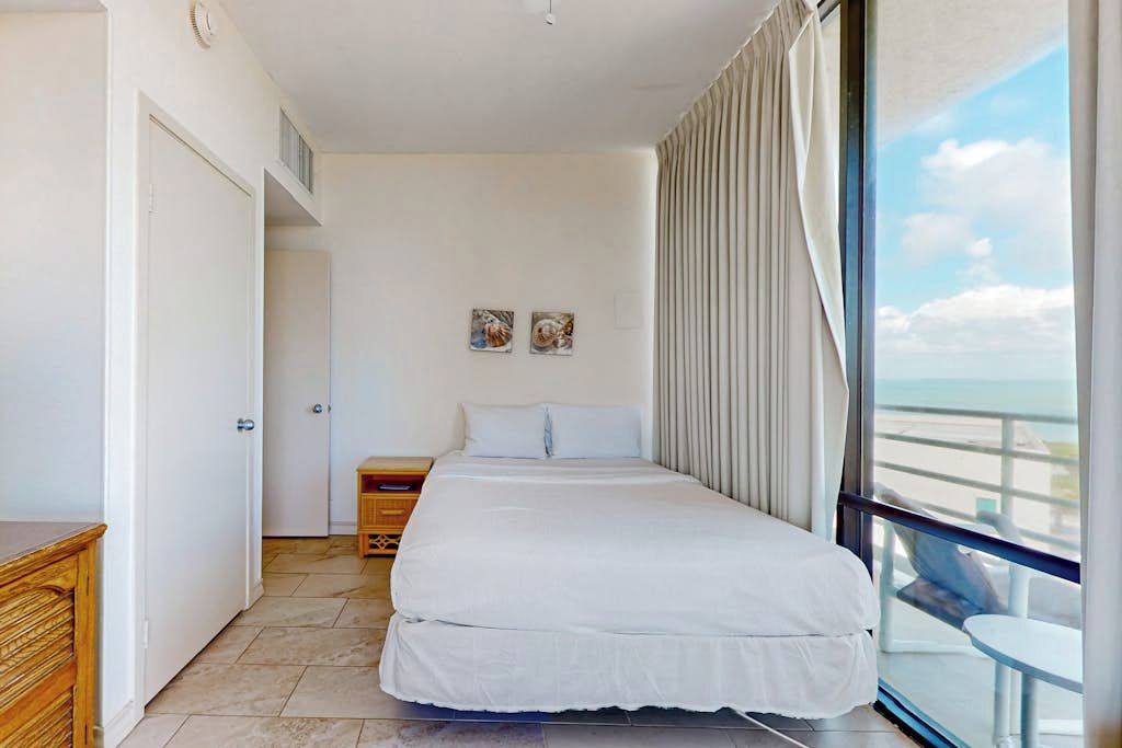 Un dormitorio con cama, mesita de noche, ventana con cortinas y vista al cielo.
