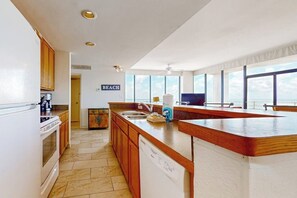 A kitchen with wooden cabinets, a white dishwasher, and a large window overlooking the sea. - Sunchase IV #715 - Gulf Views, Pools & Hot Tub (South Padre Island)