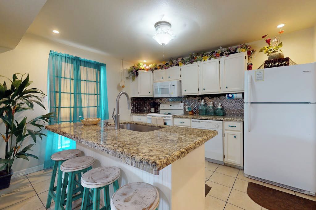 Eine Küche mit einer Granit Arbeitsplatte, weißen Schränken, einem weißen Kühlschrank und einer Spüle. Drei Barhocker stehen an der Theke.