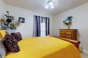 Ein Schlafzimmer mit einem Bett, einer Kommode, einem Deckenventilator, einem Fenster mit Vorhängen und einer Wanddekoration.