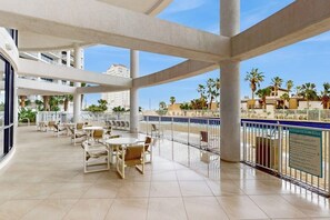 A spacious outdoor terrace with multiple levels, furnished with tables and chairs, overlooking a pool area and palm trees. - Sunchase IV #515 - Gulf & Bay Views, Hot Tub (South Padre Island)