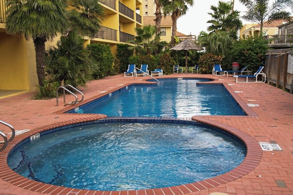 Ein rechteckiger Pool mit Backsteinumrandung, umgeben von Liegestühlen und Palmen.