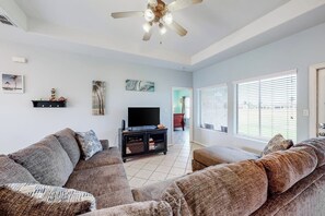 Un salon comprenant un grand canapé d’angle, un téléviseur à écran plat posé sur un meuble noir et un ventilateur de plafond.