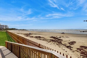 Ein Strand mit einer Holzpromenade, die zum Ufer führt, bewölkter Himmel und eine entfernte Skyline einer Stadt im Hintergrund.