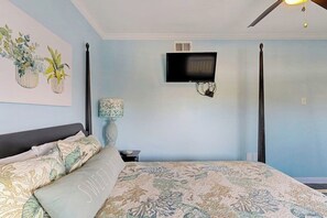 Ein Schlafzimmer mit einem Bett, einem an der Wand befestigten Fernseher, einem Deckenventilator und einem kleinen Tisch mit einer Lampe.