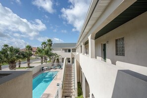 A pool area with a clear sky, palm trees, and a building with a covered walkway. - Rain Dancer Condominiums 10 (South Padre Island)