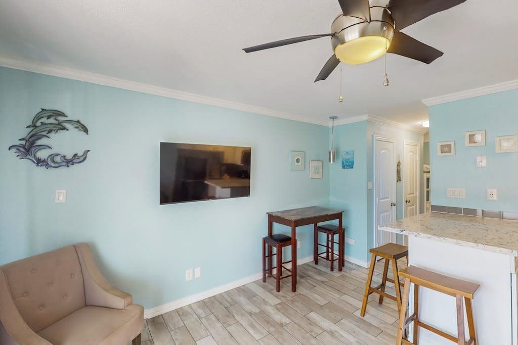 A room with a ceiling fan, a sofa, a dining table with stools, and a kitchen area with a countertop.