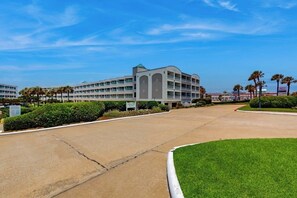 A multi-story hotel with a central courtyard, surrounded by manicured lawns and palm trees. - Seawall Getaway - Pool, Balcony, Walk to Beach (Galveston)