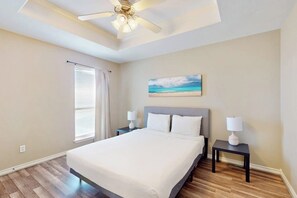 A bedroom with a bed, two bedside tables, a ceiling fan, a window with blinds, and a painting of a beach scene. - The Blue House by Whitecap Beach (Corpus Christi)