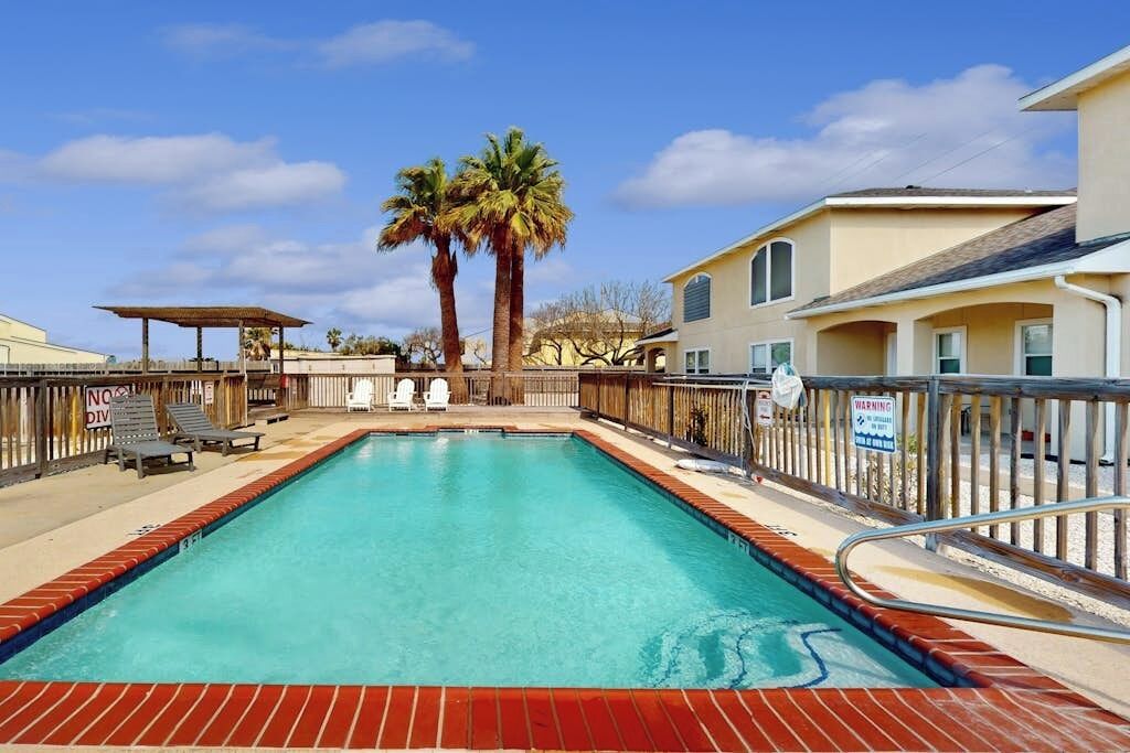 A swimming pool with a wooden deck, lounge chairs, and palm trees.