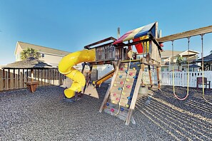 Un parc infantil amb tobogan i paret d'escalada, tot tancat per una tanca de fusta i cases al fons.