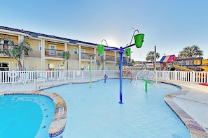 Ein Schwimmbecken mit Wasserrutsche, umgeben von einem weißen Zaun, und eine bunte Spielanlage im Hintergrund.