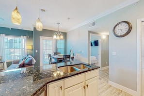 A kitchen with a granite countertop, stainless steel sink, pendant lighting, and a wall clock. - Whitecap Retreat - Steps to Beach, Pool & Balcony (Corpus Christi)