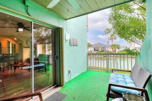 A covered outdoor area with a sliding glass door leading to a room, a green artificial turf floor, a cushioned bench, and a view of a residential area with houses and a fence. - Puente Vista 147 - Waterfront, Pool & Dock (Corpus Christi)