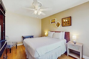 Ein Hotelzimmer mit Bett, Schreibtisch, Sessel, Nachttisch, Deckenventilator und an der Wand befestigter Kunst.