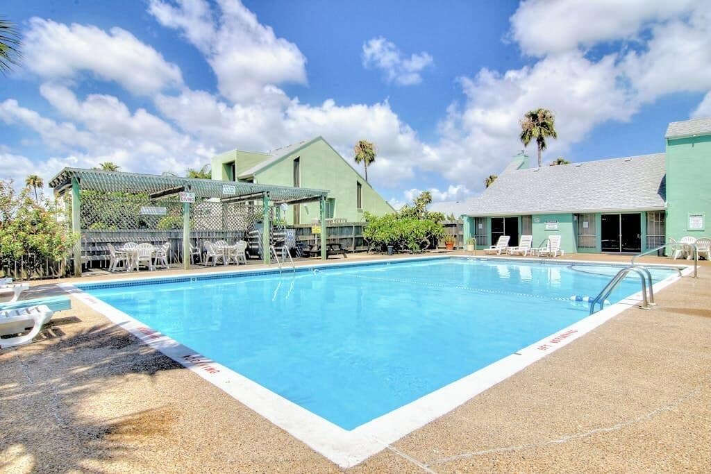 A swimming pool with lounge chairs and a covered seating area, surrounded by buildings with a tropical theme.