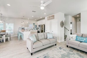 Uma cozinha moderna com armários brancos, uma área de jantar com mesa e cadeiras, uma sala de estar com sofá e almofadas decorativas, e um ventilador de teto.