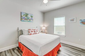 Quarto com cama, mesa de cabeceira, ventoinha no teto, janela com persianas e quadros nas paredes.