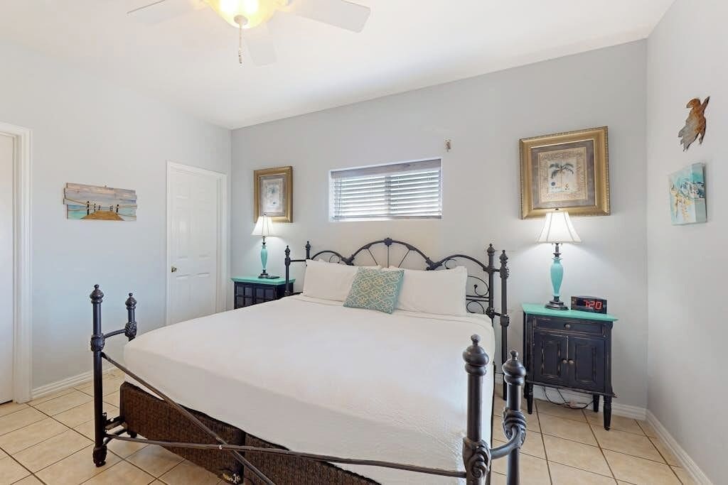 A bedroom with a four-poster bed, two bedside tables with lamps, a window with blinds, and wall art.