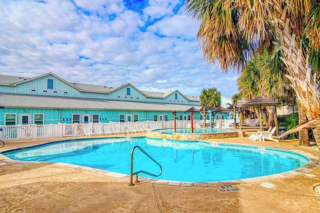 A swimming pool with a white fence, palm trees, and a building with a blue facade.