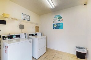 A laundry room with two washing machines, a drying rack, a trash bin, and a sign promoting easier laundry. - Quaint Oasis - Shared Pool & Beach Access (Corpus Christi)