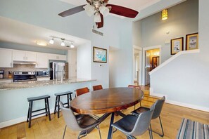 A modern kitchen with a central island, dining area with a round table, and a staircase leading to an upper level. - Lost Colony Seahorse Villa (Port Aransas)