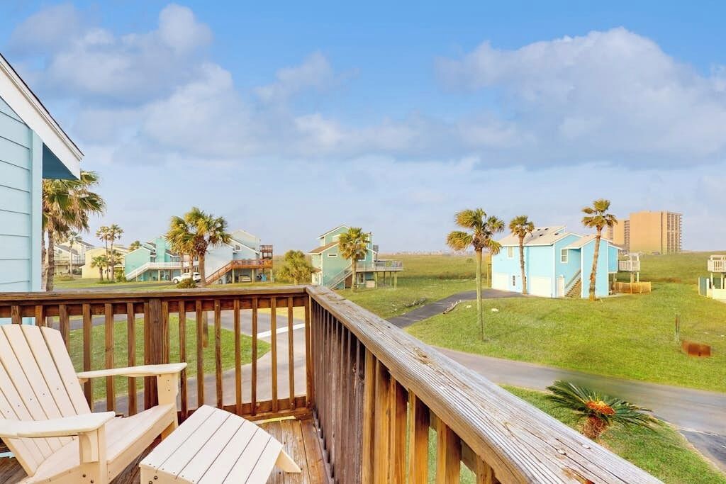 A wooden deck with chairs, a view of a coastal area with houses and palm trees, and a clear sky.
