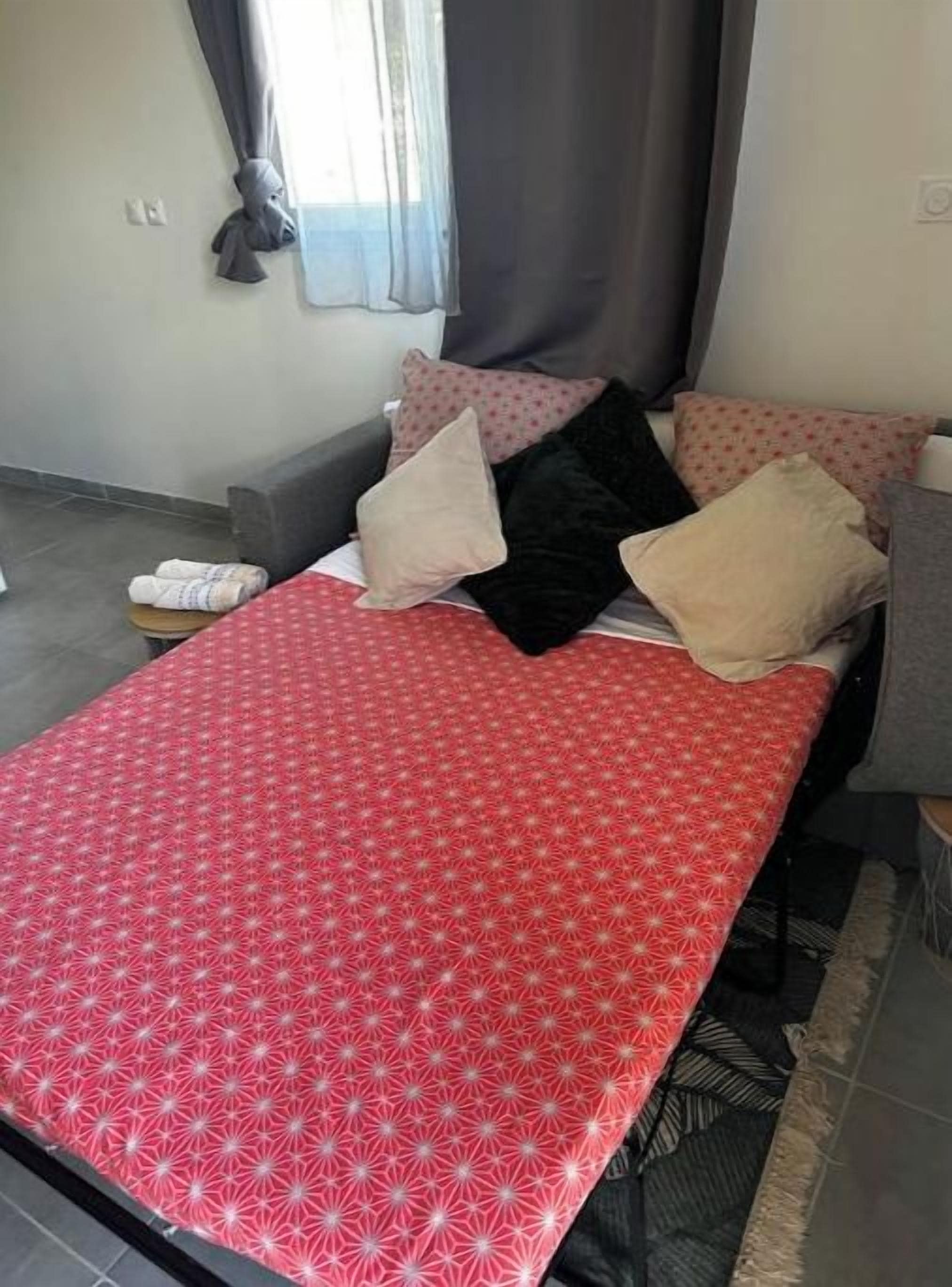 Una cama con colcha estampada, un sofá gris con almohadas y una ventana con cortinas.
