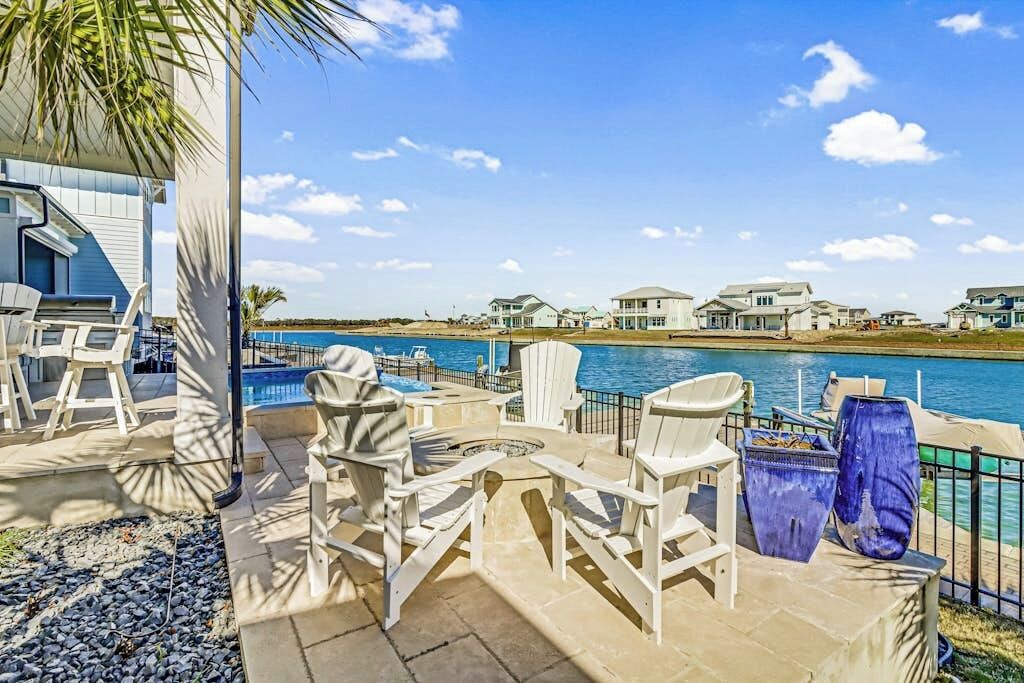Une terrasse extérieure avec des fauteuils Adirondack et un foyer, offrant une vue sur un plan d’eau et une rangée de maisons.