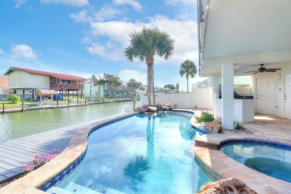A swimming pool with a wooden deck, a palm tree, and a building with a red roof in the background.