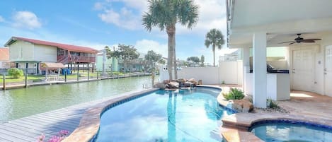 A swimming pool with a wooden deck, a palm tree, and a building with a red roof in the background.