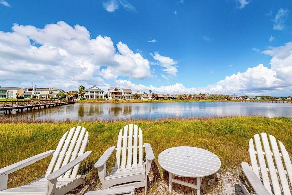 A lakeside view with three Adirondack chairs and a small table, a dock extending into the water, and houses in the background.