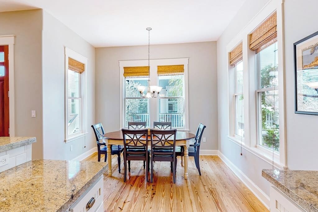 Un comedor con mesa y sillas de madera, ventanas grandes con persianas y una lámpara de araña.