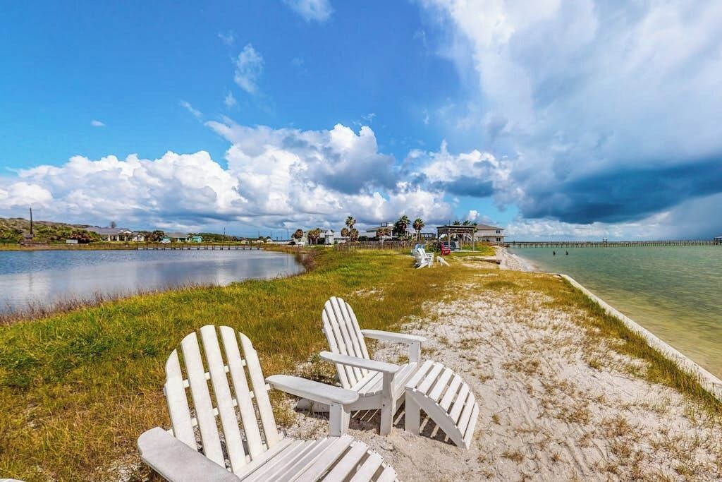 A beach with white Adirondack chairs, a pier extending into the water, and a building in the background.
