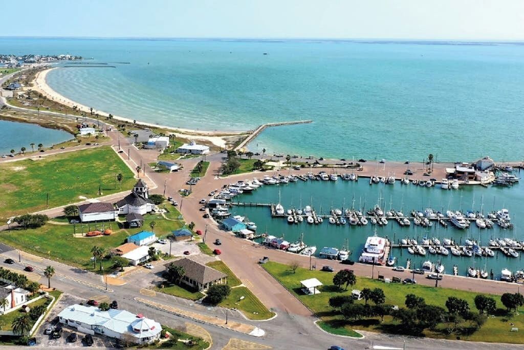 Une vue aérienne d’un port de plaisance avec des bateaux amarrés, une route menant à un bâtiment surmonté d’un dôme, et une plage en arrière-plan.