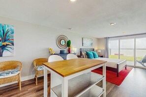 A modern living room with a wooden dining table, wicker chairs, a sofa, and a bed with a red rug. - Salt Lake Lodge - Bayfront, Big Decks & Yard (Rockport)