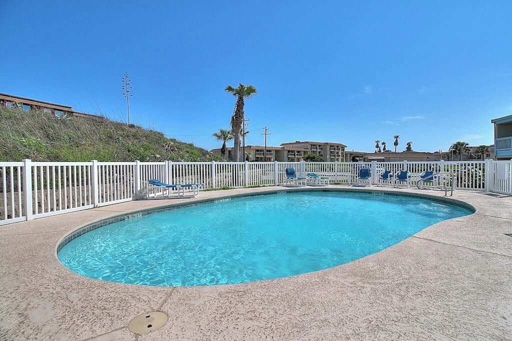 A swimming pool with a white fence, lounge chairs, and a building in the background.