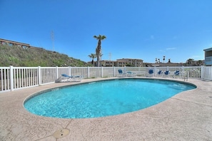 A swimming pool with a white fence, lounge chairs, and a building in the background. - Nelray Shores at Seamist Condominiums (Port Aransas)