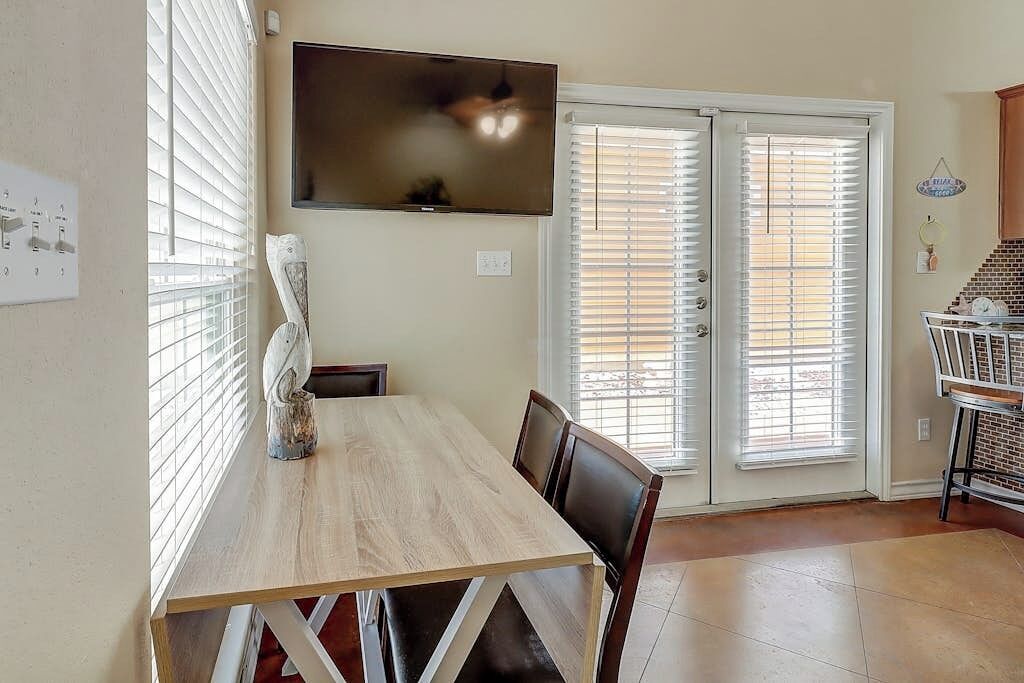 A dining area with a wooden table, chairs, a television mounted on the wall, and a sliding glass door with blinds.