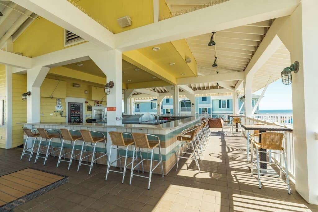 A covered outdoor bar area with multiple seating options, including bar stools and tables, and a view of the sea in the background.