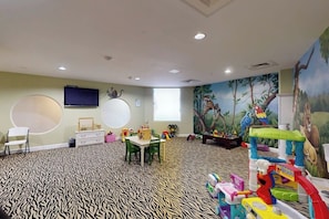 A children's playroom with a patterned carpet, a television, a mural, and various toys. - Diamond Beach Resort Seawall Stunner (Galveston)