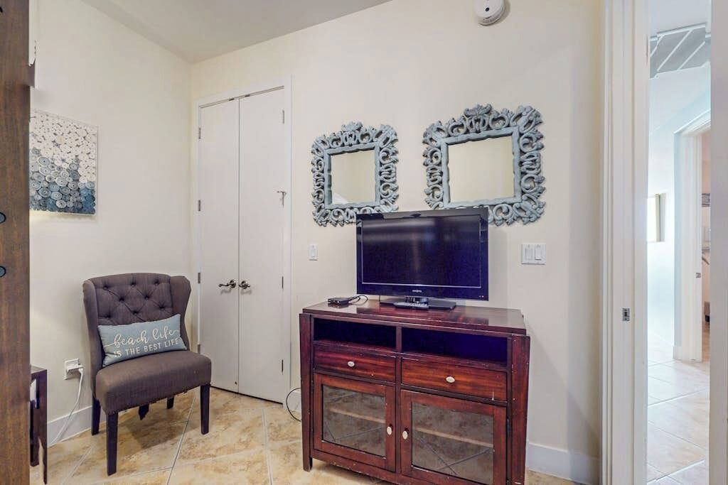 Una habitación con una televisión sobre un mueble de madera, una silla con cojín y dos espejos montados en la pared.