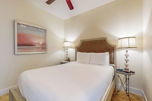 A hotel room with a large bed, two bedside tables with lamps, a wall-mounted painting, and a ceiling fan. - Diamond Beach Resort Seawall Stunner (Galveston)